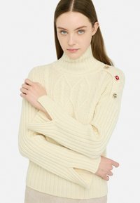 Maglione lavorato a maglia color crema con collo alto, texture a coste e motivi a treccia. Presenta bottoni decorativi in oro, rosso e accenti trasparenti.