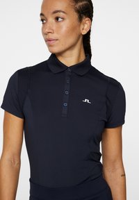 Polo bleu marine avec une patte de boutonnage, des manches courtes et des accents texturés sur les épaules. Présente un petit logo blanc sur la poitrine.