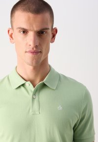 Grünes Polohemd aus Strickstoff mit strukturiertem Finish, ausgestattet mit einem Kragen und zwei weißen Knöpfen sowie einem kleinen gestickten Logo.