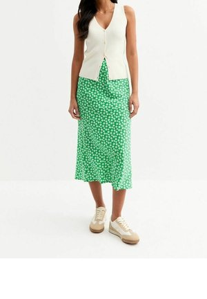 Frau trägt eine weiße ärmellose Knopfweste, einen grünen Midirock mit weißem Blumenmuster und weiße Sneakers mit beigen Sohlen.