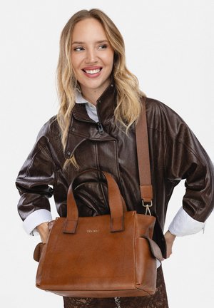 Lachende vrouw in een donkerbruine leren jas houdt een grote bruine leren handtas vast met handvatten en een schouderriem.