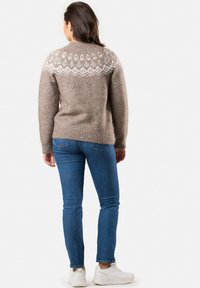 Maglione lavorato a maglia marrone con motivo geometrico bianco sulla parte superiore della schiena, abbinato a jeans blu e sneakers bianche. Texture morbida, vestibilità rilassata.