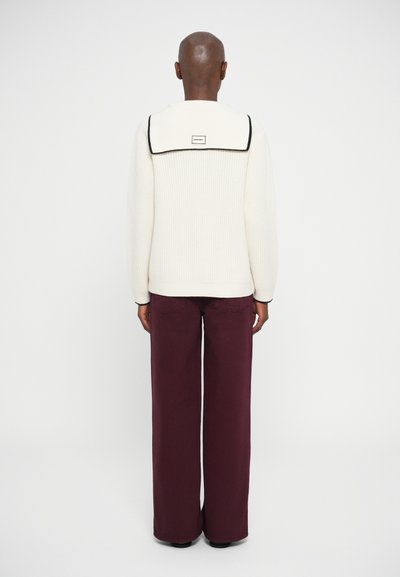 Pulover crem cu torsade și guler mare, având margini negre, asociat cu pantaloni largi de culoare burgundă. Textură netedă, design casual.