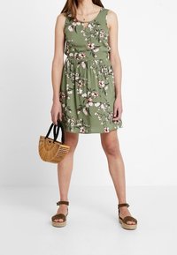 Vestido floral verde con un cuerpo ajustado y cintura elástica. Combinado con un bolso de madera marrón y sandalias beige con tiras en el tobillo.