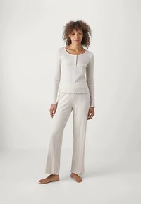 Žena stoji bosa u kremastom rebrastom dugom rukavu i odgovarajućim širim hlačama na bijeloj pozadini.