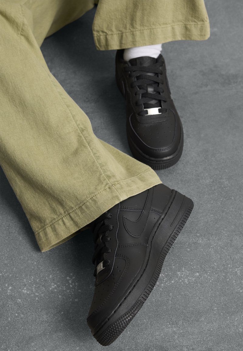 Personne portant des baskets en cuir noir, des chaussettes blanches et un pantalon vert ample, debout sur un sol gris texturé.