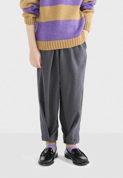 Maglione lavorato a maglia con strisce dorate e viola, pantaloni larghi grigi, mocassini neri e calze a strisce viola e dorate.