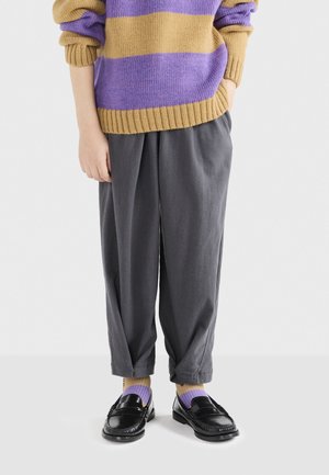 Genähter Pullover mit horizontalen lila und senfgelben Streifen, kombiniert mit grauen, lässig sitzenden Hosen und schwarzen Slippern über lila und gelben Socken.