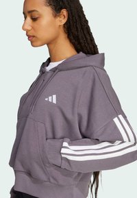 Harmaa huppari, jossa on valkoiset raidat hihoissa, etutasku ja logo rinnassa; valmistettu pehmeästä, teksturoidusta materiaalista.