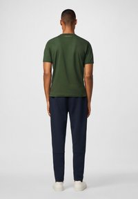 Camiseta verde de manga corta vista desde la espalda, con un cuello redondo y un sutil detalle del logo arriba, combinada con joggers azul marino y zapatillas blancas.
