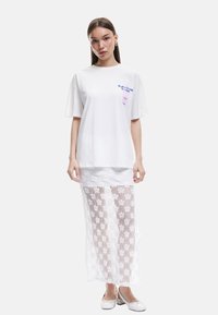 Weißes Baumwoll-T-Shirt mit kurzen Ärmeln und blauen Textdesign, kombiniert mit einem langen, weißen Spitzenrock mit floralen Mustern und transparenter Textur.