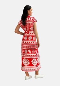Vestido largo rojo con patrones florales y geométricos en blanco, mangas cortas, cintura ceñida y dobladillo recto. Combinado con zapatillas blancas.