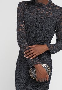 Person som bär en långärmad svart blommig spetsklänning och håller en liten svart och gyllene geometrisk väska i midjehöjd.