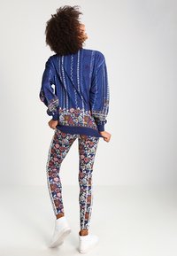 Marinblå bomberjacka med blommig fåll, matchad med mönstrade leggings i marinblått, rött och vitt. Klara ränder längs sidorna och vita sneakers.