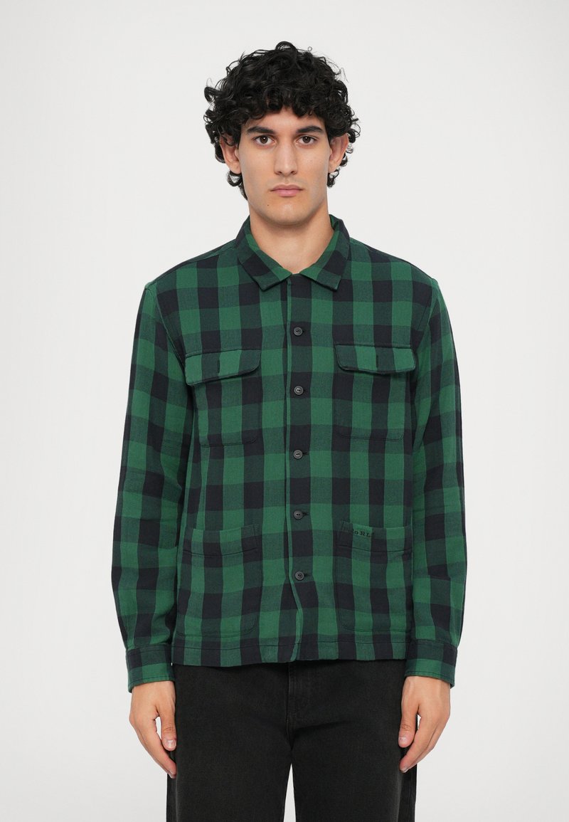 Camisa de botão xadrez verde e preta com colarinho, dois bolsos no peito e mangas longas, feita de um tecido suave e texturizado.
