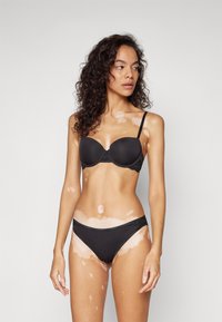 Czarny stanik z koronkowymi akcentami i wyprofilowanymi miseczkami, w zestawie z czarnymi bikini. Modelka ma kręcone włosy i skórę z jasnymi plamami.