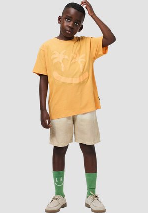 Camiseta amarilla con gráfico de palmera y cara sonriente, combinada con pantalones cortos beige claro, calcetines verdes con diseño de cara sonriente y zapatos claros.