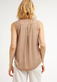 Blouse beige sans manches avec une texture douce, présentant un pli dans le dos et un ourlet arrondi. Design simple sans ornements ni motifs.