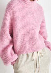 Kvinna klädd i en mjuk, texturerad rosa tröja med långa ärmar och hög krage, kombinerad med krämfärgade byxor med dragsko.