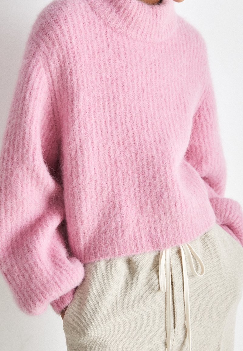 Kvinna klädd i en mjuk, texturerad rosa tröja med långa ärmar och hög krage, kombinerad med krämfärgade byxor med dragsko.