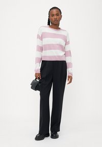 Gestreifter Strickpullover in Hellrosa und Weiß, kombiniert mit lockeren schwarzen Hosen. Das Model hält eine kleine schwarze Umhängetasche.