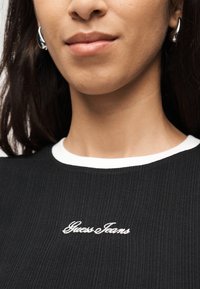 Κοντινό πλάνο ενός ατόμου που φοράει ασημένια σκουλαρίκια κρίκους και μαύρο μπλουζάκι με ρίγες και άσπρη μπορντούρα, με το λογότυπο "Guess Jeans" στο στήθος.