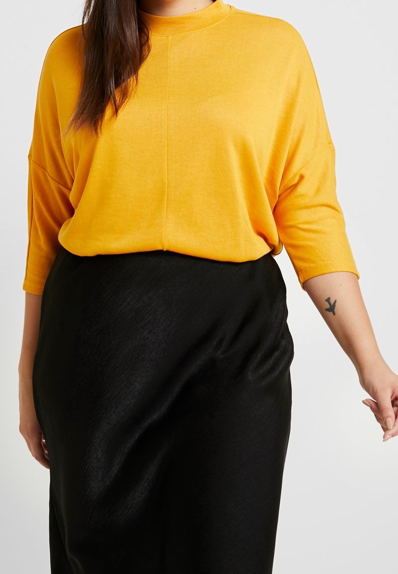 Suéter amarillo de gran tamaño con un cuello alto y mangas tres cuartos, combinado con una falda negra, elegante y de corte midi. Detalles de tela texturizada.