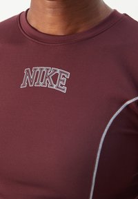 Burgundin urheilusärkki, jossa on valkoinen Nike-logo rinnassa, muotoiltu malli ja kontrastiväriset valkoiset saumat sivuilla.