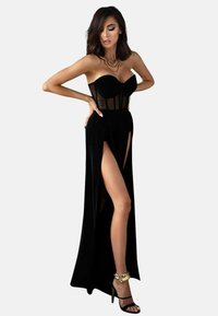 Schwarzes Abendkleid mit trägerlosem Design, durchsichtigen Bodice-Panels, hohen Seitenschlitzen und einem fließenden Rock. Kombiniert mit schwarzen High-Heels.