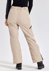 Beige geïsoleerde broek met een rekbare tailleband, zwarte ritszakken en een losse pasvorm, voorzien van ritssluitingen bij de enkels voor eenvoudig aan- en uittrekken.