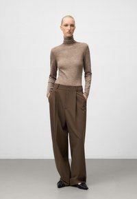 Brązowy sweter z golfem zestawiony z brązowymi spodniami o wysokim stanie i szerokich nogawkach. Model pozuje z rękami w kieszeniach, prezentując czarne buty wsuwane.