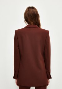Bruine op maat gemaakte blazer met scherpe revers, gestructureerde schouders en lange mouwen. Vloeiende stof met een schone achterkant en subtiele naaddetails.