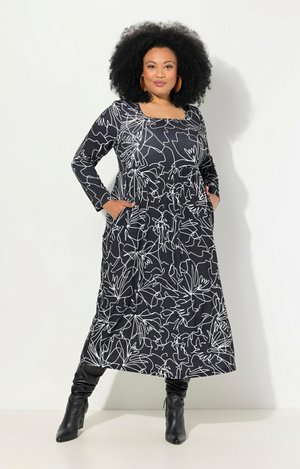 Schwarzes langärmliges Kleid mit weißen blumigen Linienmustern, runder Halsauschnitt und seitlichen Taschen. Kombiniert mit schwarzen knielangen Stiefeln.