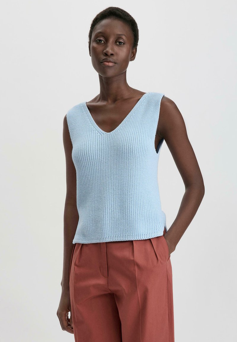 Lichtblauwe gebreide mouwloze top met een V-hals, textuurstof en een rechte zoom, gecombineerd met high-waisted roestkleurige broek.