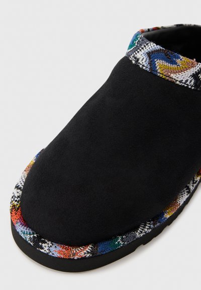 Zapatilla de ante negro con un ribete de tejido decorado que presenta diseños zigzag multicolores. Punta redondeada y suela negra flexible.