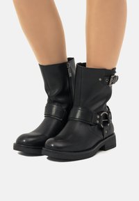 Botas de cuero negro hasta el tobillo con cierre de cremallera, correa ancha con hebilla de plata, punta redondeada y suela texturizada.