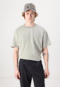Ljusgrön t-shirt med kort ärm och texturerad yta, kombinerad med mörkblå byxor. Grå keps prydd med färgglada broderade lappar.