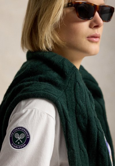 Personne blonde portant des lunettes de soleil écaille de tortue, un polo blanc de Wimbledon avec un logo sur la manche, et un pull en maille torsadée vert foncé posé sur les épaules.
