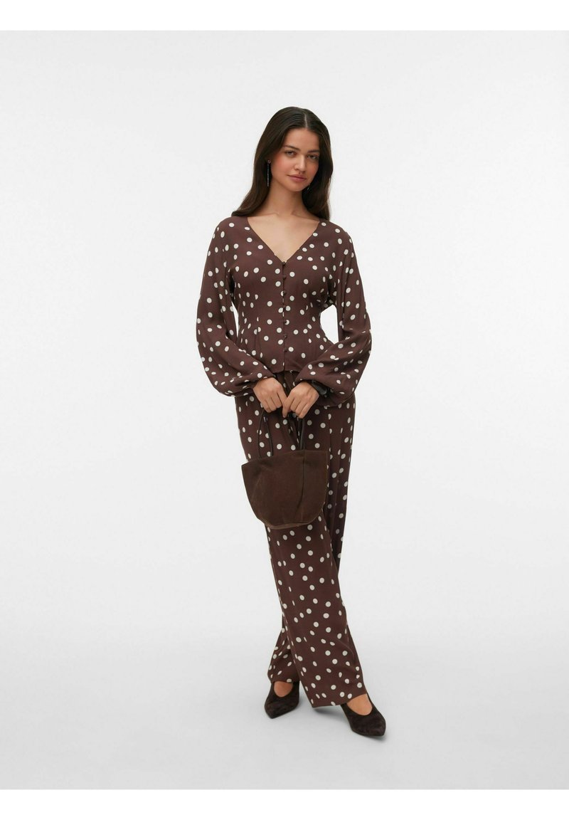 Femme portant un chemisier marron à pois et un pantalon, tenant un sac à main marron assorti, debout devant un fond blanc.