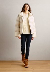 Világos krémszínű puff Jacket, krémszínű kötött pulóver, sötét farmernadrág és barna, fehér talpú, prémes csizmák.