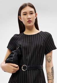 Jauna moteris su tiesiais rudais plaukais, dėvinti juodą dryžuotą suknelę su juodu diržu, laikanti juodą tekstūruotą rankinę ir rodanti tatuiruotes ant rankų.