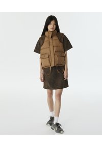 Gewatteerd bruin vest met hoge kraag, voorzien van twee grote zakken aan de voorkant; gedragen over een gestructureerde donkerbruine jurk met spikkels. Grijze sneakers met witte sokken.