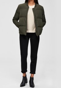 Giacca a puff verde oliva con scollo rotondo, abbinata a un maglione chiaro e pantaloni neri risvoltati. Stivaletti neri completano il look.