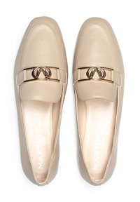 Mocasines de cuero color beige con un acabado suave, que presentan un acento de doble logo en tono dorado en la parte delantera y un diseño de punta redondeada.
