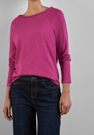 Osoba ubrana w jaskraworóżowy top z długim rękawem i okrągłym dekoltem oraz ciemnoniebieskie dżinsy na tle jednolitego szarego tła.