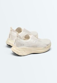 Paire de chaussures de sport beiges sans lacets avec semelles épaisses et rembourrées et tissu en maille, inclinées pour montrer les détails latéraux et arrière.