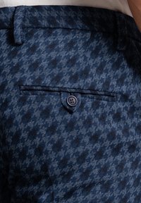 Kalhoty námořnicky modré barvy s vzorem kostičky, viditelná textura látky. Mají zadní kapsu se zapínáním na tmavě modré tlačítko.