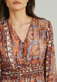 Langärmliges Kleid mit einem aufwendigen orange, blauen und braunen Paisley-Muster, V-Ausschnitt und einer gerafften Taille mit Rüschensaum.