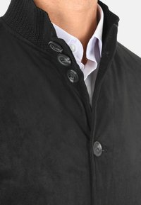 Giacca in suede nero con colletto a coste, dotata di tre bottoni, una texture liscia e una camicia bianca visibile sotto.