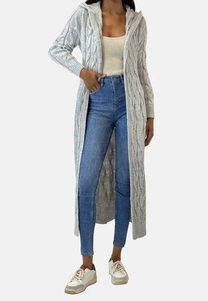 Femme portant un cardigan long gris clair en maille torsadée, un débardeur beige, un jean skinny taille haute bleu, et des baskets blanches à lacets.
